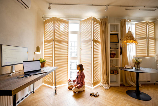 Young woman meditates after work, sitting on the floor at cozy home office. Stylish interior with workplace and computers in front. Mental health and relaxation, working from home concept - Φωτογραφία, εικόνα
