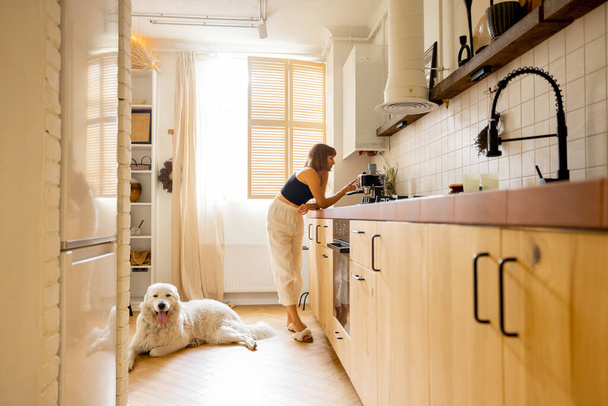 Женщина варит кофе, стоя со своей собакой на кухне. Интерьер современной и стильной кухни в бежевых тонах. Концепция бытового образа жизни - Фото, изображение