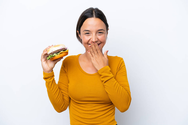 Jovem hispânica segurando um hambúrguer isolado no fundo branco feliz e sorrindo cobrindo a boca com a mão - Foto, Imagem