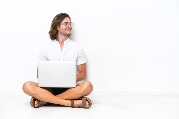 Fiatal jóképű férfi egy laptop ül a padlón elszigetelt fehér háttér látszó oldalán - Fotó, kép