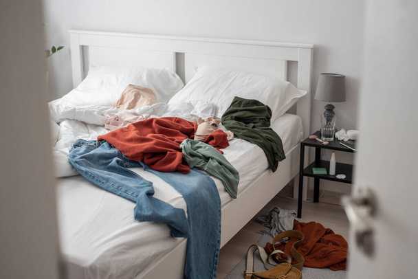 View to the cluttered bedroom through a half open door. Clothes thrown all around the bed. Cluttered bedroom with a white bedding and a minimalistic modern interior. - Fotó, kép