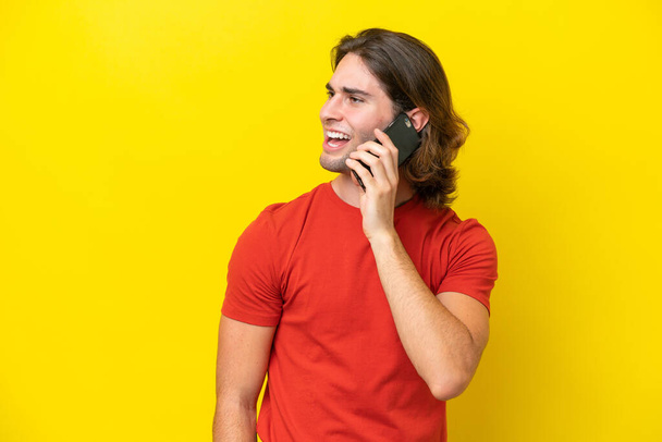 Белый красивый мужчина, изолированный на желтом фоне, поддерживает разговор с кем-то по мобильному телефону - Фото, изображение