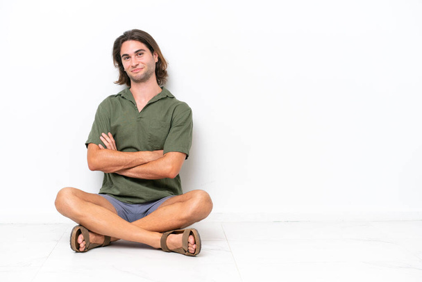 Jovem homem bonito sentado no chão isolado no fundo branco com os braços cruzados e olhando para a frente - Foto, Imagem