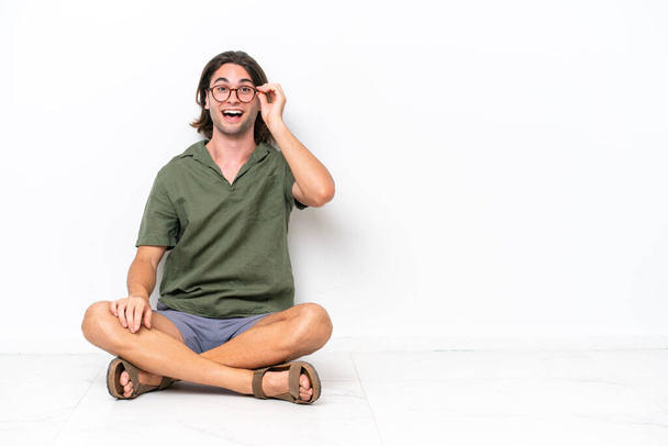 Νεαρός όμορφος άνδρας κάθεται στο πάτωμα απομονωμένος σε λευκό φόντο με γυαλιά και έκπληκτος - Φωτογραφία, εικόνα