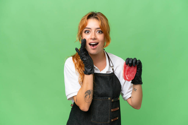 Χασάπης καυκάσιος γυναίκα φοράει ποδιά και σερβίρει φρέσκο κομμένο κρέας απομονωμένο σε πράσινο φόντο chroma οθόνη με έκπληξη και σοκαρισμένη έκφραση του προσώπου - Φωτογραφία, εικόνα