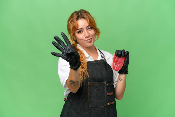 Boucher femme caucasienne portant un tablier et servir de la viande fraîche coupée isolé sur fond vert chroma clé heureux et comptant quatre avec des doigts - Photo, image