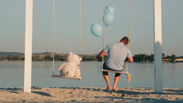 Un uomo solo insieme a un orsacchiotto e palle gonfiabili siedono su un'altalena vicino a un lago. Triste compleanno nella solitudine. Un uomo solo - Filmati, video