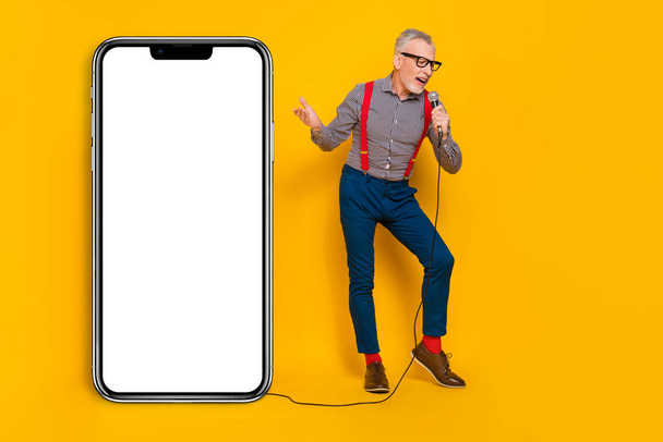 Full body photo of attractive pensioner mister hold mic sing song near phone promo banner isolated on yellow color background. - 写真・画像