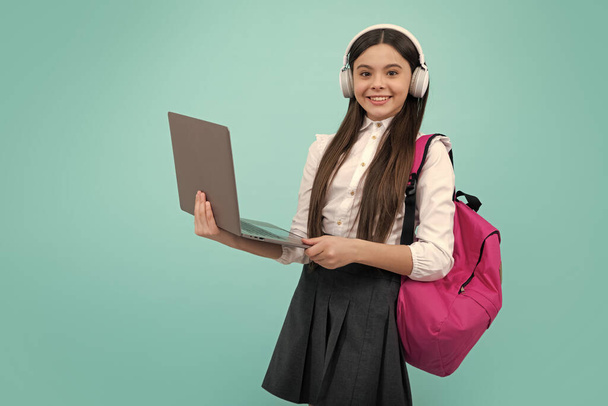 Back to school. Happy teenager school girl in school uniform with bagpack, headphones and laptop. School children on isolated blue background - Photo, Image