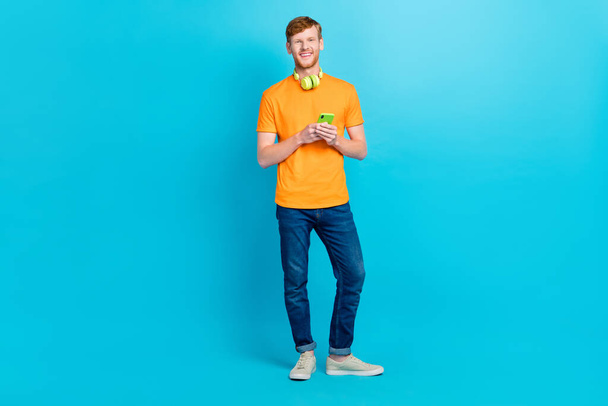 Full size photo of trend red hair guy hold telephone wear t-shirt jeans shoes isolated on teal color background. - Foto, Imagem