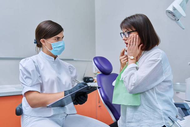 Dentist and woman patient in dental clinic, doctor nurse writing on clipboard consulting, middle aged female talking about toothache disturbing dental problem. Dentistry, treatment, dental health care - Foto, Imagem