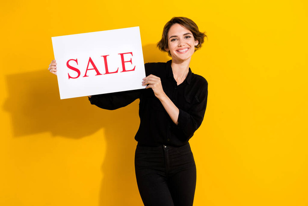 Photo of cute millennial short hair lady hold poster sale wear black shirt jeans isolated on yellow color background. - Φωτογραφία, εικόνα