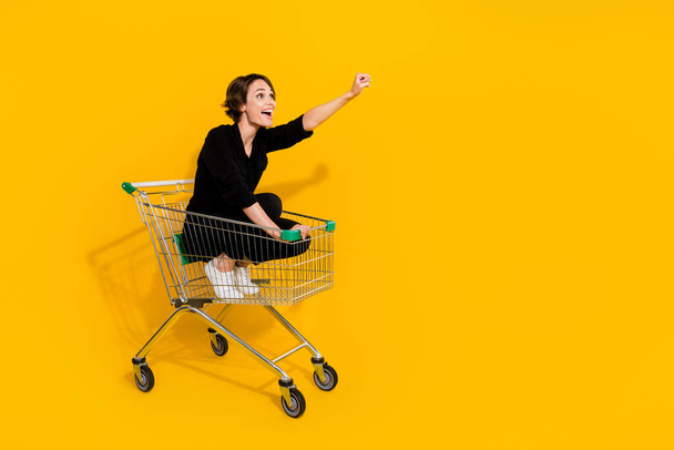Photo of sweet charming girl dressed formal shirt riding shopping tray rising fist empty space isolated yellow color background. - Foto, Imagem