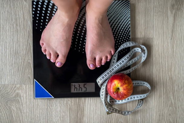 Imagem cortada de mulher em pé em escalas contra fita métrica e maçã. Envolvemos as pernas com uma fita métrica. medidas de peso corporal. pés femininos - Foto, Imagem