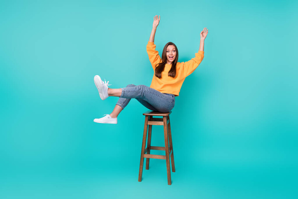 Full length photo of young adorable pretty nice woman wear yellow sweater jeans hands up celebrate party nice mood sitting bar isolated on cyan color background. - Fotoğraf, Görsel