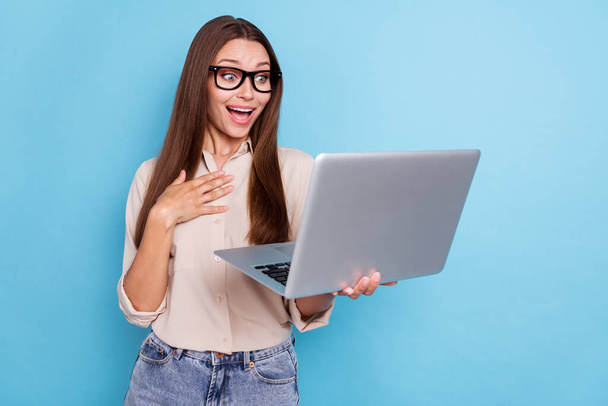 Portrait of gorgeous smart impressed girl with long hair wear beige shirt hold laptop hand on chest isolated on blue color background. - 写真・画像