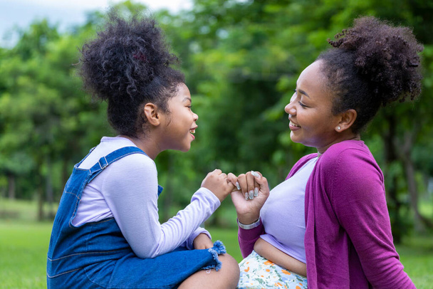 Afroamerikanische Mutter gibt ihrer kleinen Tochter beim sommerlichen Picknick im öffentlichen Park ein pinkfarbenes Versprechen für Liebe und Glück - Foto, Bild