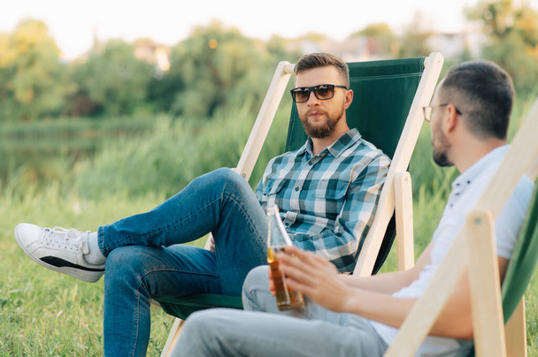 Two men sitting in camping chairs drinking beer and chatting outdoors. - Photo, image