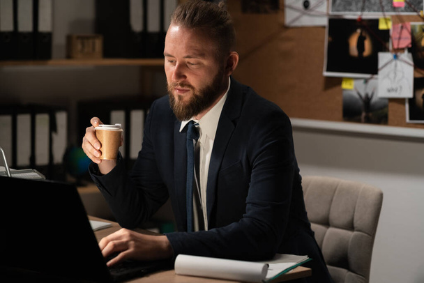 Detektiv arbeitet im Büro mit einem Computer, Prozess der Analyse der Beweise für einen Kriminalfall, Ermittler trinkt Kaffee in der Nacht am Arbeitsplatz - Foto, Bild