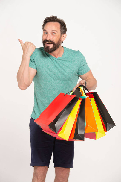 Bearded smiling man holding shopping bags and pointing by thumb up to copy space over white background and looking at camera - Fotó, kép