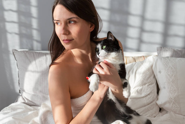 Portret van een mooi meisje met een kat. Slaapkamerachtergrond. Het meisje knuffelt de kat en kijkt weg. Ochtend in bed. - Foto, afbeelding