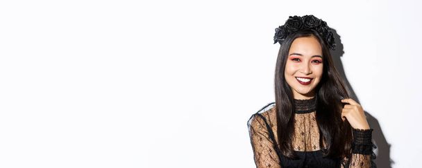 Primer plano de hermosa mujer asiática elegante en corona negra y vestido de encaje gótico sonriendo, de pie sobre fondo blanco, vestido para la fiesta de halloween. - Foto, imagen