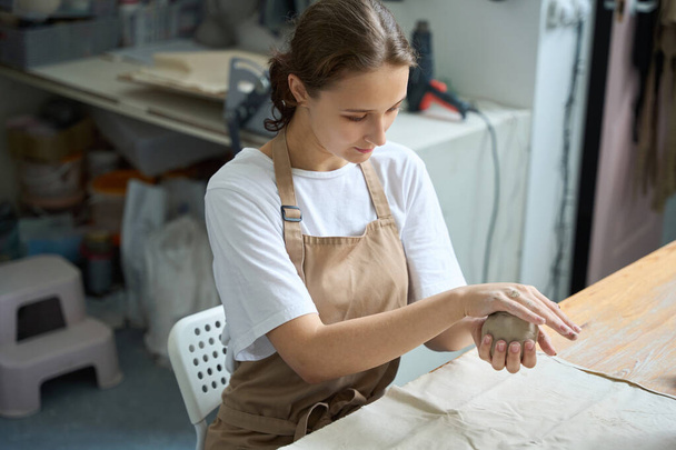 Mujer joven tranquila trabajando cuidadosamente con un bulto redondo de arcilla en un aula - Foto, imagen