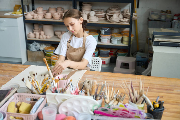 Young woman in a special apron sculpts clay at her desk in a spacious pottery studio - 写真・画像