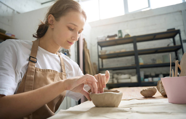 明るい工房の作業台の若い女性は、慎重に粘土板を彫刻し、彼女の手は粘土で染色されています - 写真・画像
