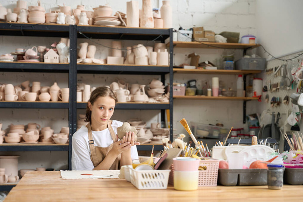 Beautiful craftswoman sits table with piece of clay in her hands against the background of a rack with clay blanks - 写真・画像