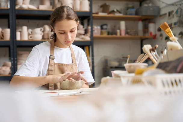 Mujer joven en el estudio de cerámica trabaja en una mesa y extiende los bordes de una arcilla en blanco con los dedos - Foto, imagen