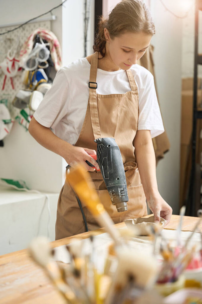 Estudiante en un delantal de protección seca el blanco de una placa de arcilla con un secador de pelo especial en un estudio de cerámica - Foto, Imagen