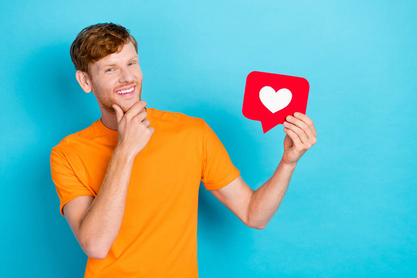 Photo of positive clever man wear orange t-shirt arm chin holding heart like card isolated teal color background. - Фото, зображення