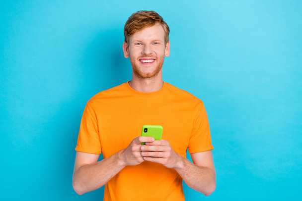 Photo of handsome toothy beaming guy with ginger hairstyle dressed orange t-shirt hold smartphone isolated on teal color background. - Fotó, kép