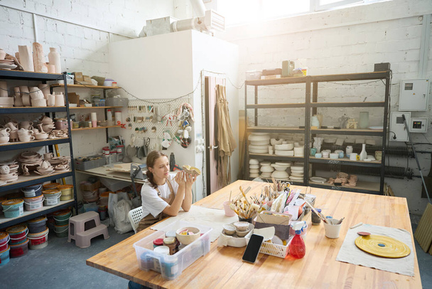 Nice woman from a bright studio at a large wooden table paints a homemade clay plate - Photo, Image