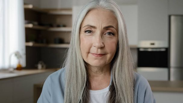 Portrait calm confident adult middle-aged woman pensioner granny mistress homeowner tender mature old lady with long grey hair posing in kitchen room serene looking at camera healthy beauty appearance - 写真・画像