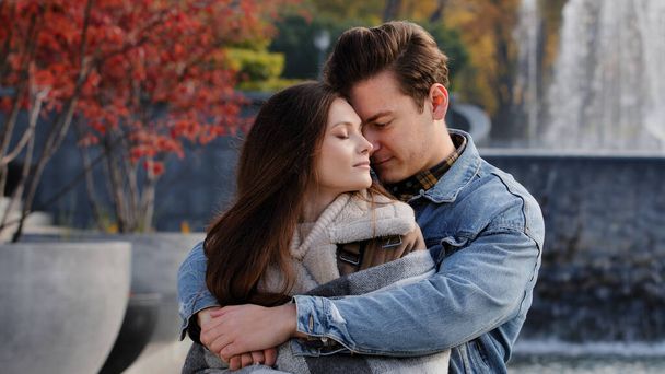 Caucasian couple lovers partners stand background of city fountain pressed closely to cheeks together spend time in autumn park on date embrace feel love emotion romantic atmosphere happy young family - Photo, Image