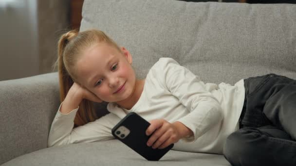 O usuário esperto do miúdo da menina júnior da geração nova usa o descanso moderno do smartphone no bate-papo do sofá mensagens de texto em linha através do jogo do messenger social jogos vídeo móveis conecte-se ao wifi viciado à criança do gadget - Filmagem, Vídeo