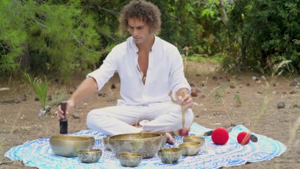 A man playing on a tibetian singing bowls sitting outdoors for meditation. Buddhist healing practices. Sound therapy, alternative medicine. Clearing the space of negative energy. Selective focus - Footage, Video