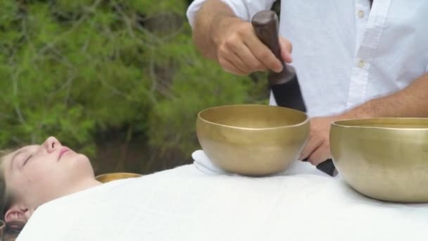 A therapist of sound massage therapy doing a Buddhist healing practices for a young woman laying on a massage bed with tibetan singing bowls outdoor. Spiritual practic for harmony, balance, wellbeing - Footage, Video