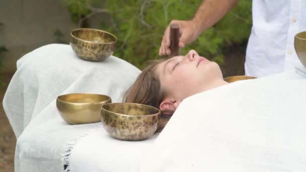 Un terapeuta de la terapia de masaje de sonido haciendo prácticas budistas de curación para una mujer joven acostada en una cama de masaje con cuencos de canto tibetano al aire libre. Práctica espiritual para armonía, equilibrio, bienestar - Imágenes, Vídeo