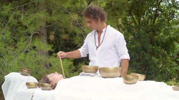 A man wearing in white doing healing sound massage with tibetan singing bowl to a woman, practice sound therapy with Nepal Buddha singing bowl outdoor, young woman relax with alternative medicine - Footage, Video