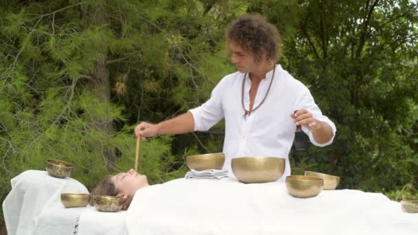 A man wearing in white doing healing sound massage with tibetan singing bowl to a woman, practice sound therapy with Nepal Buddha singing bowl outdoor, young woman relax with alternative medicine - Footage, Video