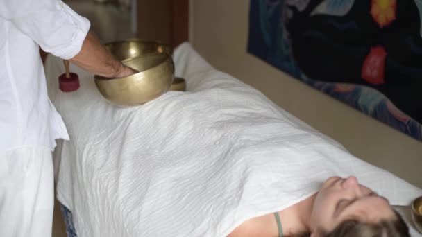 Un uomo che indossa in bianco facendo massaggio sonoro curativo con tibetano canto ciotola ad una donna, pratica la terapia del suono con Nepal Buddha canto ciotola all'aperto, giovane donna rilassarsi con la medicina alternativa - Filmati, video