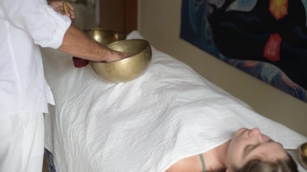 Un uomo che indossa in bianco facendo massaggio sonoro curativo con tibetano canto ciotola ad una donna, pratica la terapia del suono con Nepal Buddha canto ciotola all'aperto, giovane donna rilassarsi con la medicina alternativa - Filmati, video