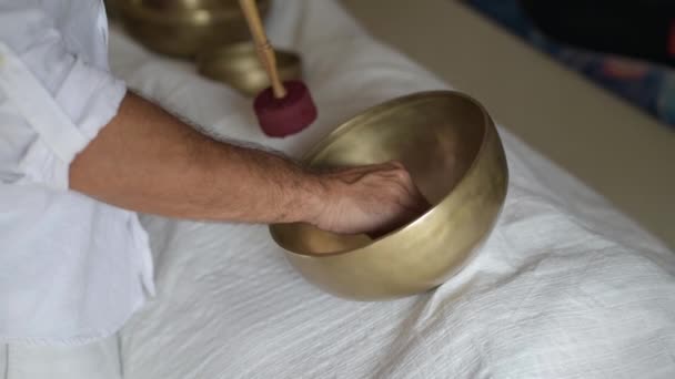 Ein Therapeut der Klangmassagetherapie praktiziert eine buddhistische Heilpraxis für eine junge Frau, die auf einem Massagebett mit tibetischen Klangschalen im Innenraum liegt. Spirituelle Praxis für Harmonie, Gleichgewicht, Wohlbefinden - Filmmaterial, Video