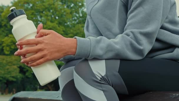 Close up unknown athlete healthy woman runner wear sporting suit sit on bench in park rest after training hard workout exercises take break pause hold sports bottle of fresh filtered water relax alone - Footage, Video