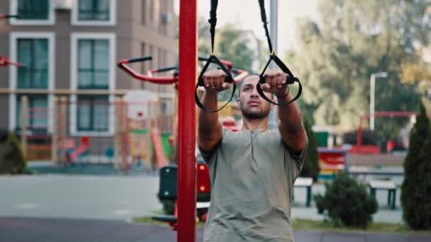 Erkek vücut geliştirici atletik Latin adam yazın spor sahasında antrenman yapıyor askıya alınmış TRX kayışlarıyla egzersiz yapıyor kaslarını esnetiyor ve sokak sporu yapıyor. - Video, Çekim
