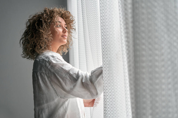Zijaanzicht van dromerige tedere vrouw met krullend haar in wit shirt en wegkijkend in het raam met wit gordijn bij zacht daglicht - Foto, afbeelding