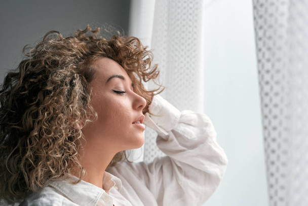 Side view of romantic young female with curls wearing white shirt keeping eyes closed while touching hair standing near window in daylight - Foto, Imagem